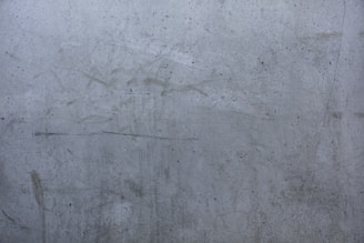 A close-up shot of a rough, gray concrete wall with numerous scratches, stains, and small dark spots. The wall appears weathered and aged.