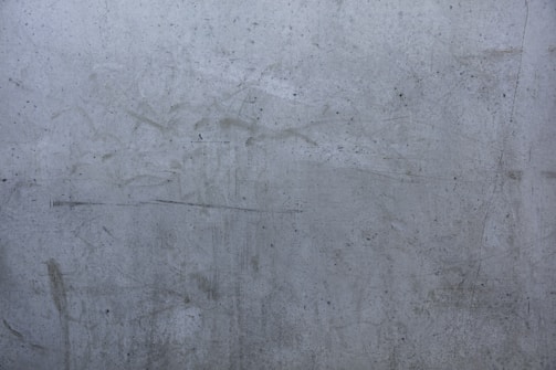 A close-up shot of a rough, gray concrete wall with numerous scratches, stains, and small dark spots. The wall appears weathered and aged.