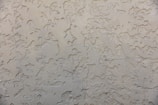A textured wall surface with a rough, irregular pattern. The surface appears to be made of a beige or light tan material and is uniformly covered with a plaster-like texture, creating a rustic and aged appearance.