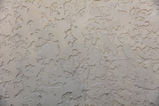 A textured wall surface with a rough, irregular pattern. The surface appears to be made of a beige or light tan material and is uniformly covered with a plaster-like texture, creating a rustic and aged appearance.