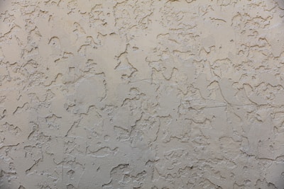 A textured wall surface with a rough, irregular pattern. The surface appears to be made of a beige or light tan material and is uniformly covered with a plaster-like texture, creating a rustic and aged appearance.