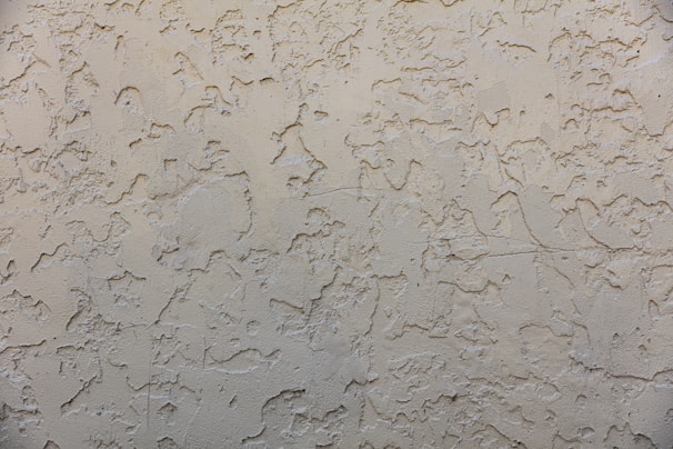 A textured wall surface with a rough, irregular pattern. The surface appears to be made of a beige or light tan material and is uniformly covered with a plaster-like texture, creating a rustic and aged appearance.
