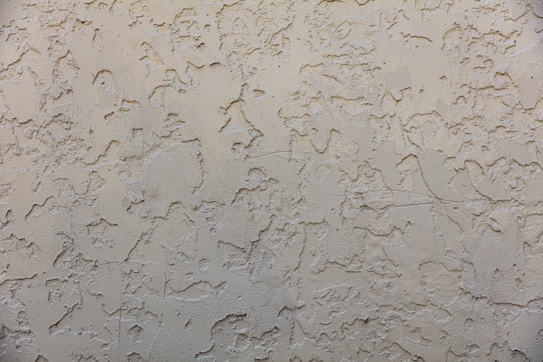 A textured wall surface with a rough, irregular pattern. The surface appears to be made of a beige or light tan material and is uniformly covered with a plaster-like texture, creating a rustic and aged appearance.