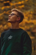 A young person with short, light brown hair wearing glasses and a dark sweatshirt featuring a space-themed design stands against a blurred background of warm, golden foliage. They are looking upward, creating a sense of contemplation.