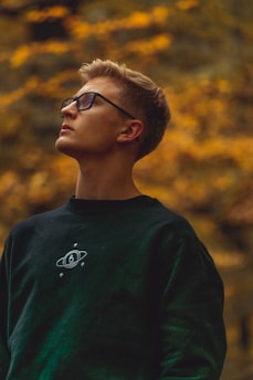 A young person with short, light brown hair wearing glasses and a dark sweatshirt featuring a space-themed design stands against a blurred background of warm, golden foliage. They are looking upward, creating a sense of contemplation.