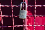 A metallic padlock with a keyhole marked 'BEST' hangs on a grid of metallic wires painted in a reddish hue. The background features a blurred red surface providing a vibrant contrast to the lock.