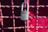 A metallic padlock with a keyhole marked 'BEST' hangs on a grid of metallic wires painted in a reddish hue. The background features a blurred red surface providing a vibrant contrast to the lock.