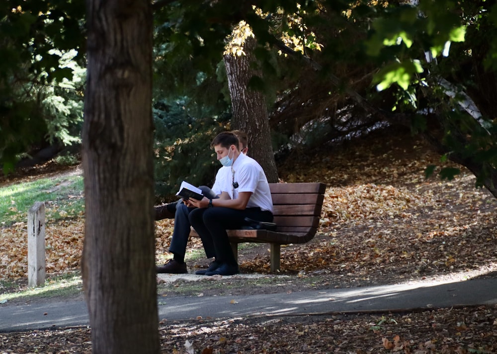 분홍색 드레스 셔츠를 입은 남자가 갈색 나무 벤치에 앉아 책을 읽고 있다