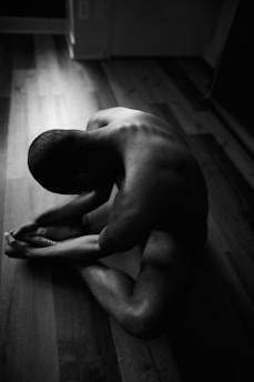 A person seated on a hardwood floor in a dimly lit space, showing a muscular back and limbs in an introspective pose. The lighting emphasizes the contours of the body, creating strong contrasts between light and shadow.