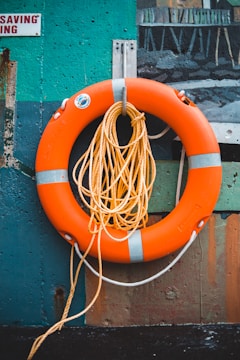 A bright orange life preserver with white stripes is mounted against a colorful, textured wall. A coiled yellow rope is draped around the life preserver, adding to the nautical theme of the scene.