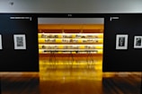 An art gallery with framed black and white photographs displayed on walls and a central glowing yellow-lit shelf. The floor is shiny and wooden, with a table centered in front of the illuminated display. The walls have names of cities labeled on them.