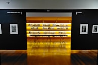 An art gallery with framed black and white photographs displayed on walls and a central glowing yellow-lit shelf. The floor is shiny and wooden, with a table centered in front of the illuminated display. The walls have names of cities labeled on them.