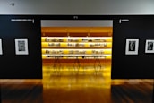 An art gallery with framed black and white photographs displayed on walls and a central glowing yellow-lit shelf. The floor is shiny and wooden, with a table centered in front of the illuminated display. The walls have names of cities labeled on them.