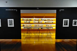 An art gallery with framed black and white photographs displayed on walls and a central glowing yellow-lit shelf. The floor is shiny and wooden, with a table centered in front of the illuminated display. The walls have names of cities labeled on them.