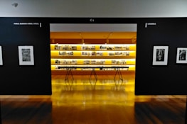 An art gallery with framed black and white photographs displayed on walls and a central glowing yellow-lit shelf. The floor is shiny and wooden, with a table centered in front of the illuminated display. The walls have names of cities labeled on them.
