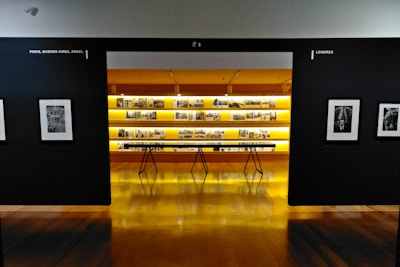 An art gallery with framed black and white photographs displayed on walls and a central glowing yellow-lit shelf. The floor is shiny and wooden, with a table centered in front of the illuminated display. The walls have names of cities labeled on them.