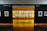 An art gallery with framed black and white photographs displayed on walls and a central glowing yellow-lit shelf. The floor is shiny and wooden, with a table centered in front of the illuminated display. The walls have names of cities labeled on them.