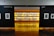 An art gallery with framed black and white photographs displayed on walls and a central glowing yellow-lit shelf. The floor is shiny and wooden, with a table centered in front of the illuminated display. The walls have names of cities labeled on them.