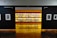 An art gallery with framed black and white photographs displayed on walls and a central glowing yellow-lit shelf. The floor is shiny and wooden, with a table centered in front of the illuminated display. The walls have names of cities labeled on them.