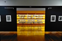 An art gallery with framed black and white photographs displayed on walls and a central glowing yellow-lit shelf. The floor is shiny and wooden, with a table centered in front of the illuminated display. The walls have names of cities labeled on them.