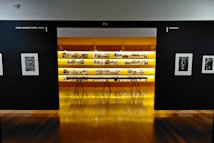 An art gallery with framed black and white photographs displayed on walls and a central glowing yellow-lit shelf. The floor is shiny and wooden, with a table centered in front of the illuminated display. The walls have names of cities labeled on them.