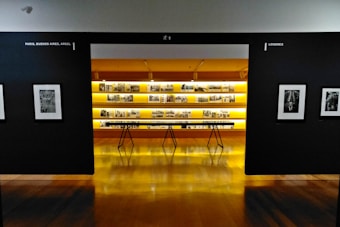 An art gallery with framed black and white photographs displayed on walls and a central glowing yellow-lit shelf. The floor is shiny and wooden, with a table centered in front of the illuminated display. The walls have names of cities labeled on them.