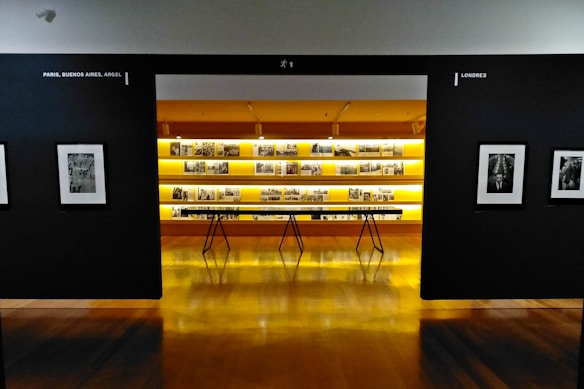 An art gallery with framed black and white photographs displayed on walls and a central glowing yellow-lit shelf. The floor is shiny and wooden, with a table centered in front of the illuminated display. The walls have names of cities labeled on them.