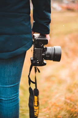 A person dressed in a dark jacket and jeans is holding a professional camera with a brand strap. The background consists of a softly blurred field with autumnal colors, suggesting an outdoor setting.