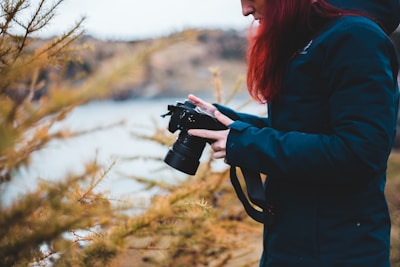 A person dressed in a dark jacket holds a camera surrounded by branches with golden leaves. The setting appears outdoors, with a blurred natural landscape in the background, suggesting a focus on the photographic activity.