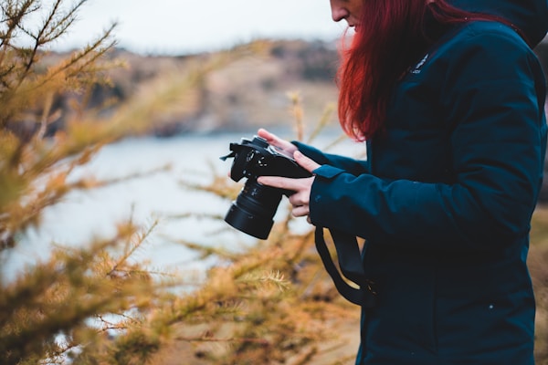 A person dressed in a dark jacket holds a camera surrounded by branches with golden leaves. The setting appears outdoors, with a blurred natural landscape in the background, suggesting a focus on the photographic activity.