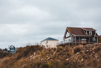 A rural landscape with two houses, one with a pitched roof and large windows poised on a rocky, grassy hilltop under an overcast sky.