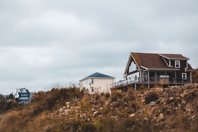 A rural landscape with two houses, one with a pitched roof and large windows poised on a rocky, grassy hilltop under an overcast sky.