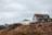 A rural landscape with two houses, one with a pitched roof and large windows poised on a rocky, grassy hilltop under an overcast sky.