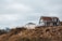 A rural landscape with two houses, one with a pitched roof and large windows poised on a rocky, grassy hilltop under an overcast sky.