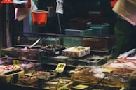 A busy seafood market stall displaying various fresh seafood items on ice. The stall features transparent tanks and containers filled with shellfish, fish, and other sea creatures. There are handwritten price tags visible, and bright lighting contrasts with the dark background.