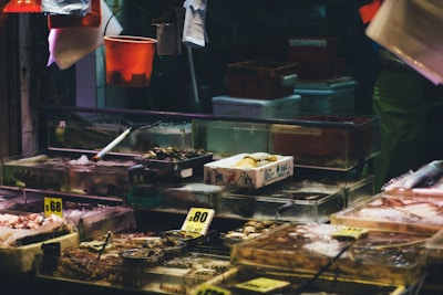 A busy seafood market stall displaying various fresh seafood items on ice. The stall features transparent tanks and containers filled with shellfish, fish, and other sea creatures. There are handwritten price tags visible, and bright lighting contrasts with the dark background.