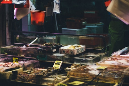 A busy seafood market stall displaying various fresh seafood items on ice. The stall features transparent tanks and containers filled with shellfish, fish, and other sea creatures. There are handwritten price tags visible, and bright lighting contrasts with the dark background.