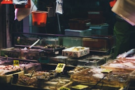 A busy seafood market stall displaying various fresh seafood items on ice. The stall features transparent tanks and containers filled with shellfish, fish, and other sea creatures. There are handwritten price tags visible, and bright lighting contrasts with the dark background.
