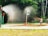 A playground scene with water sprinklers creating a misty environment. Three children are playing near the sprinklers, with one child in a red outfit standing directly under the spray. The ground is wet with water, and lush green trees form a dense backdrop.