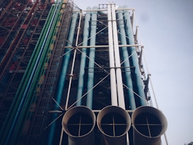 A large, industrial structure featuring multiple large, colorful pipes running vertically alongside a metal framework. The pipes are predominantly green, blue, and white, with some structural elements in gray. The composition is complex and suggests a piece of modern architecture or an industrial facility.