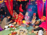 A traditional wedding ceremony with colorful attire and decorations. Several people, including a bride and groom, are seated around a ritual fire. The scene is vibrant with red drapery and floral arrangements, creating a festive atmosphere.