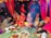 A traditional wedding ceremony with colorful attire and decorations. Several people, including a bride and groom, are seated around a ritual fire. The scene is vibrant with red drapery and floral arrangements, creating a festive atmosphere.