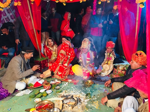 A traditional wedding ceremony with colorful attire and decorations. Several people, including a bride and groom, are seated around a ritual fire. The scene is vibrant with red drapery and floral arrangements, creating a festive atmosphere.