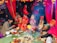 A traditional wedding ceremony with colorful attire and decorations. Several people, including a bride and groom, are seated around a ritual fire. The scene is vibrant with red drapery and floral arrangements, creating a festive atmosphere.