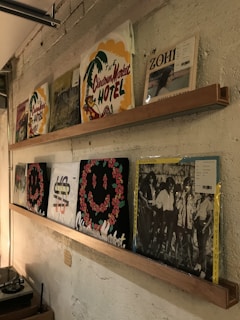 A wall with two wooden shelves displaying vinyl record covers. The covers feature various designs, including a palm tree with 'The Chinatown Market Hotel' text, a record with a floral smiley face, and black and white images of people. The background is a textured, painted wall.