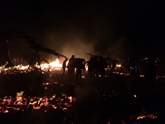 A group of people is standing near a large bonfire at night, creating silhouettes against the flames. The scene is illuminated by the warm glow of the fire, with smoke rising into the dark sky. Burned structures and charred wood are visible in the foreground.