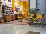 A cozy store interior with shelves filled with various colorful bottles and products. The store has a warm color palette, with yellow walls and natural wood furniture. A yellow bicycle with a basket filled with yellow flowers is parked against a brown column near a reflective floor.