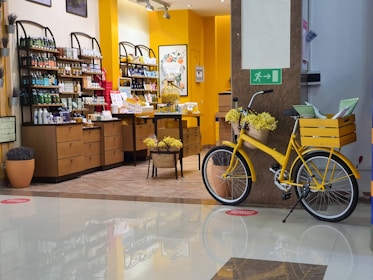 A cozy store interior with shelves filled with various colorful bottles and products. The store has a warm color palette, with yellow walls and natural wood furniture. A yellow bicycle with a basket filled with yellow flowers is parked against a brown column near a reflective floor.