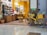 A cozy store interior with shelves filled with various colorful bottles and products. The store has a warm color palette, with yellow walls and natural wood furniture. A yellow bicycle with a basket filled with yellow flowers is parked against a brown column near a reflective floor.