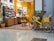 A cozy store interior with shelves filled with various colorful bottles and products. The store has a warm color palette, with yellow walls and natural wood furniture. A yellow bicycle with a basket filled with yellow flowers is parked against a brown column near a reflective floor.